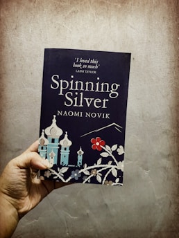 A hand holds a book titled 'Spinning Silver' by Naomi Novik. The cover features stylized illustrations of buildings with onion-shaped domes and floral designs. A quote from Laini Taylor is visible at the top.