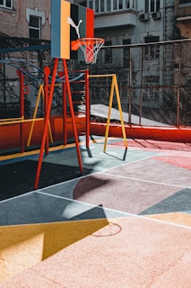 A colorful urban playground with a basketball hoop attached to a multicolored backboard. The ground is painted with geometric patterns, and there is a swing set nearby. The setting is surrounded by buildings, giving it a vibrant, urban feel.
