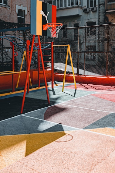 A colorful urban playground with a basketball hoop attached to a multicolored backboard. The ground is painted with geometric patterns, and there is a swing set nearby. The setting is surrounded by buildings, giving it a vibrant, urban feel.