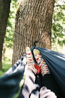 A pair of feet wearing colorful, geometric-patterned socks rest comfortably inside a hammock. The hammock is suspended between trees in a lush, green outdoor setting, suggesting a peaceful, leisurely environment.