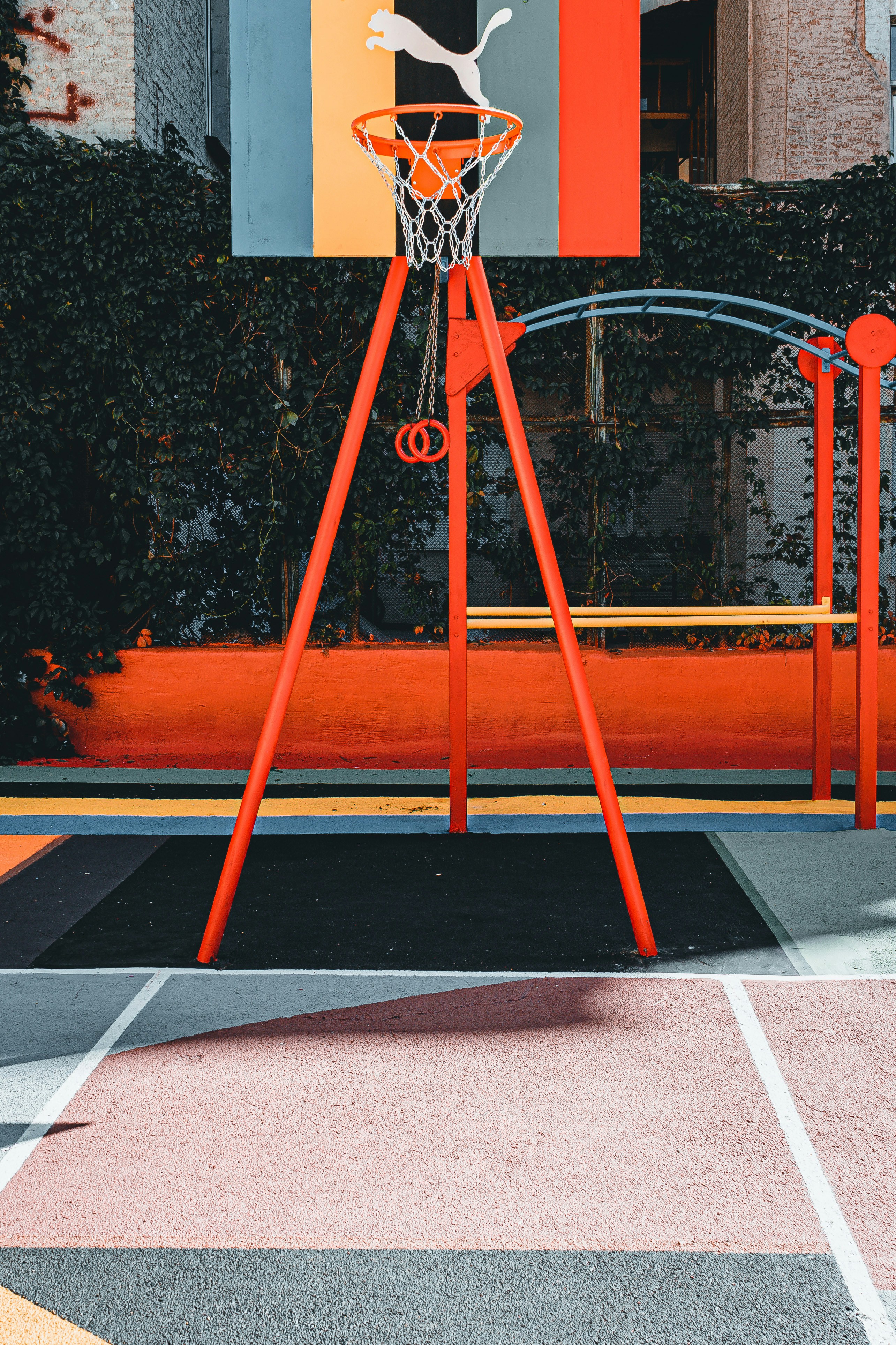 A vibrant outdoor basketball court with a red and orange theme. The basketball hoop stands prominently with a colorful backboard featuring geometric designs, situated against a background of lush greenery.
