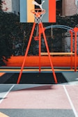 A vibrant outdoor basketball court with a red and orange theme. The basketball hoop stands prominently with a colorful backboard featuring geometric designs, situated against a background of lush greenery.