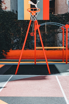 A vibrant outdoor basketball court with a red and orange theme. The basketball hoop stands prominently with a colorful backboard featuring geometric designs, situated against a background of lush greenery.