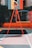 A vibrant outdoor basketball court with a red and orange theme. The basketball hoop stands prominently with a colorful backboard featuring geometric designs, situated against a background of lush greenery.