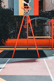 A vibrant outdoor basketball court with a red and orange theme. The basketball hoop stands prominently with a colorful backboard featuring geometric designs, situated against a background of lush greenery.