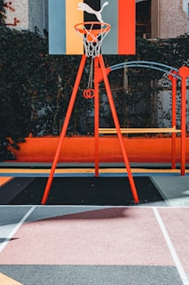 A vibrant outdoor basketball court with a red and orange theme. The basketball hoop stands prominently with a colorful backboard featuring geometric designs, situated against a background of lush greenery.