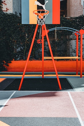 A vibrant outdoor basketball court with a red and orange theme. The basketball hoop stands prominently with a colorful backboard featuring geometric designs, situated against a background of lush greenery.