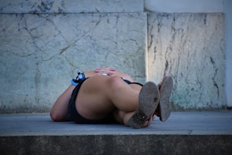 A person is lying on a stone surface outdoors with their legs crossed and hands resting on their abdomen. They are in casual clothing, and one of their wrists has a blue wristband. The background features a textured stone wall.