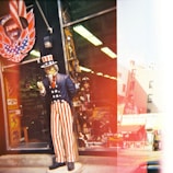 A life-sized statue of Uncle Sam stands outside a store, dressed in a blue jacket, red and white striped pants, and a top hat with stars. The store window is filled with various merchandise and decorations. There is a person inside the shop visible through the glass. The image has a vintage effect with streaks of red and white light.