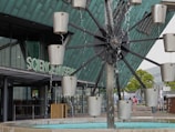 A modern architectural structure with the words Science Museum visible on the glass facade. In the foreground, a water feature with rotating metal containers creates a dynamic, kinetic sculpture. Various individuals are seen walking and observing the surroundings. The building's exterior is composed of glass and metal, giving it a contemporary and industrial look.