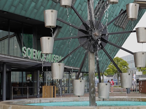 A modern architectural structure with the words Science Museum visible on the glass facade. In the foreground, a water feature with rotating metal containers creates a dynamic, kinetic sculpture. Various individuals are seen walking and observing the surroundings. The building's exterior is composed of glass and metal, giving it a contemporary and industrial look.