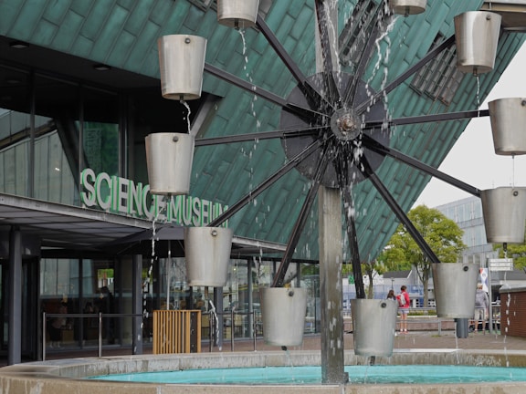 A modern architectural structure with the words Science Museum visible on the glass facade. In the foreground, a water feature with rotating metal containers creates a dynamic, kinetic sculpture. Various individuals are seen walking and observing the surroundings. The building's exterior is composed of glass and metal, giving it a contemporary and industrial look.