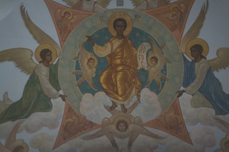 A religious painting featuring a central figure with a golden robe, seated on a cloud, holding a book or scroll. Surrounding the central figure are several winged angels. The background contains geometric shapes and clouds, contributing to a celestial atmosphere.