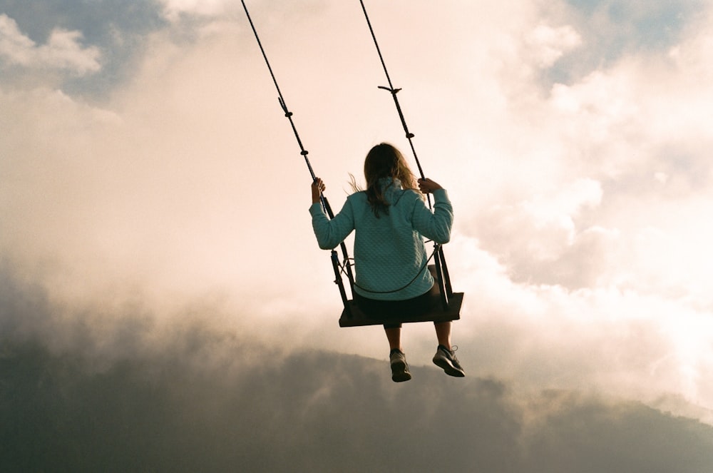fille en sweat à capuche bleu assis sur balançoire sous ciel nuageux pendant la journée
