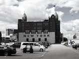 A historic, multi-story hotel building, labeled 'NEW YORK', with two distinctive towers topped with green domes and clocks. The building is situated behind a parking lot filled with various cars and a small utility vehicle under a cloudy sky.