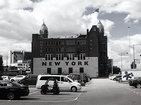 A historic, multi-story hotel building, labeled 'NEW YORK', with two distinctive towers topped with green domes and clocks. The building is situated behind a parking lot filled with various cars and a small utility vehicle under a cloudy sky.