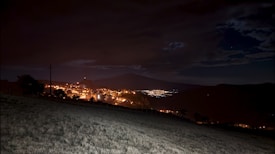 A nighttime landscape featuring a warmly lit town sprawling across a hillside. The foreground consists of a gently sloping field, while the dark silhouette of a mountain forms the backdrop. Clouds are scattered across the sky, with patches of deep blue night visible.