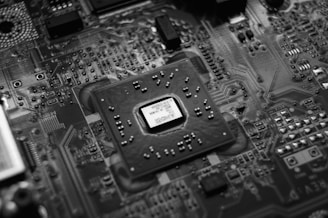 A monochrome close-up view of an electronic circuit board featuring a central microchip surrounded by various components such as resistors and capacitors. The intricate network of pathways and components displays a complex design typical of modern electronic devices.