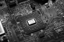 A monochrome close-up view of an electronic circuit board featuring a central microchip surrounded by various components such as resistors and capacitors. The intricate network of pathways and components displays a complex design typical of modern electronic devices.