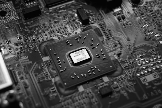 A monochrome close-up view of an electronic circuit board featuring a central microchip surrounded by various components such as resistors and capacitors. The intricate network of pathways and components displays a complex design typical of modern electronic devices.