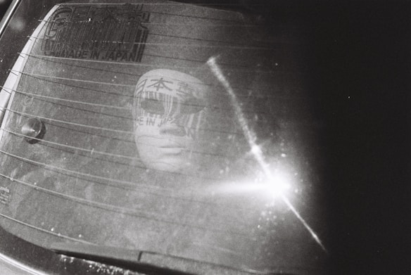 A mysterious mask-like face is reflected on the back window of a car. The window is marked with horizontal defogger lines and has stickers with Japanese writing. Sunlight creates a bright glare on the glass.