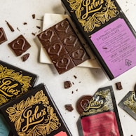 A collection of cannabis-infused chocolate bars and pieces, elegantly presented with intricate packaging designs and heart-shaped details on the chocolate. The packaging includes vibrant colors and elaborate patterns, featuring gold and black elements with distinct labeling.