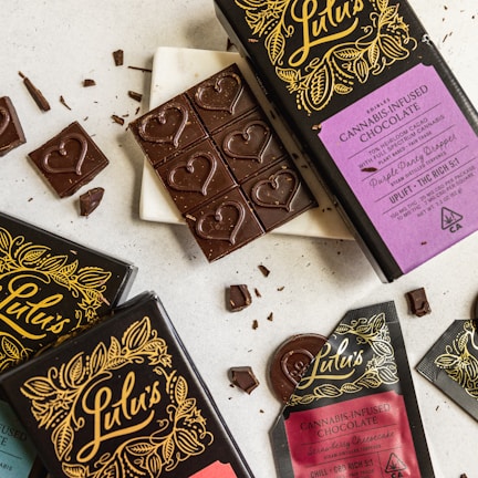 A collection of cannabis-infused chocolate bars and pieces, elegantly presented with intricate packaging designs and heart-shaped details on the chocolate. The packaging includes vibrant colors and elaborate patterns, featuring gold and black elements with distinct labeling.
