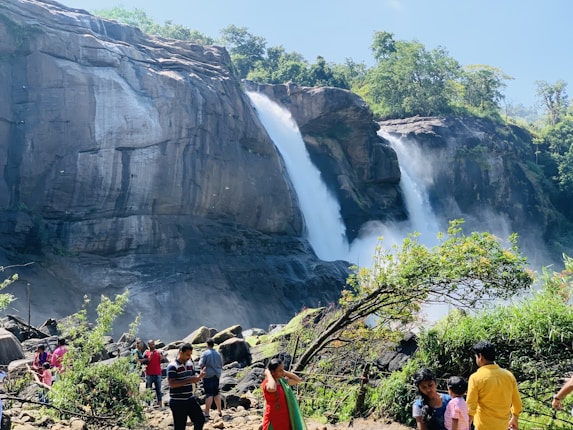 A large waterfall cascades over a rocky cliff surrounded by lush greenery. In the foreground, several people are gathered, some are taking photographs while others are simply enjoying the natural scenery. The sky is clear, adding to the brightness of the scene.