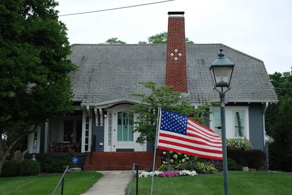 A charming house with a gray shingled roof, featuring a prominent red brick chimney. The front yard is well-maintained with green grass and colorful flowers lining the pathway. An American flag waves from a pole in the foreground, and a vintage-style streetlamp stands near the entrance. A cozy porch with seating is visible to the left of the entrance.