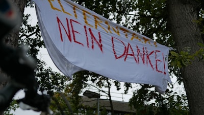 A white banner with red and yellow text hangs between two trees. The background shows green foliage and a hint of architectural structure.
