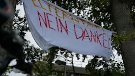 A white banner with red and yellow text hangs between two trees. The background shows green foliage and a hint of architectural structure.