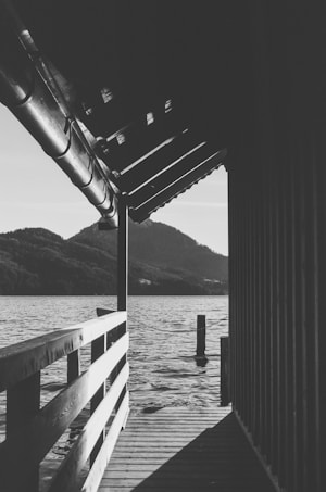 A black and white photograph captures a lakeside scene. A wooden dock with a railing extends over the water, leading toward a structure that partially covers the walkway. In the background, mountains rise beneath a clear sky. The serene surface of the lake reflects the light, contributing to the tranquil and contemplative atmosphere.