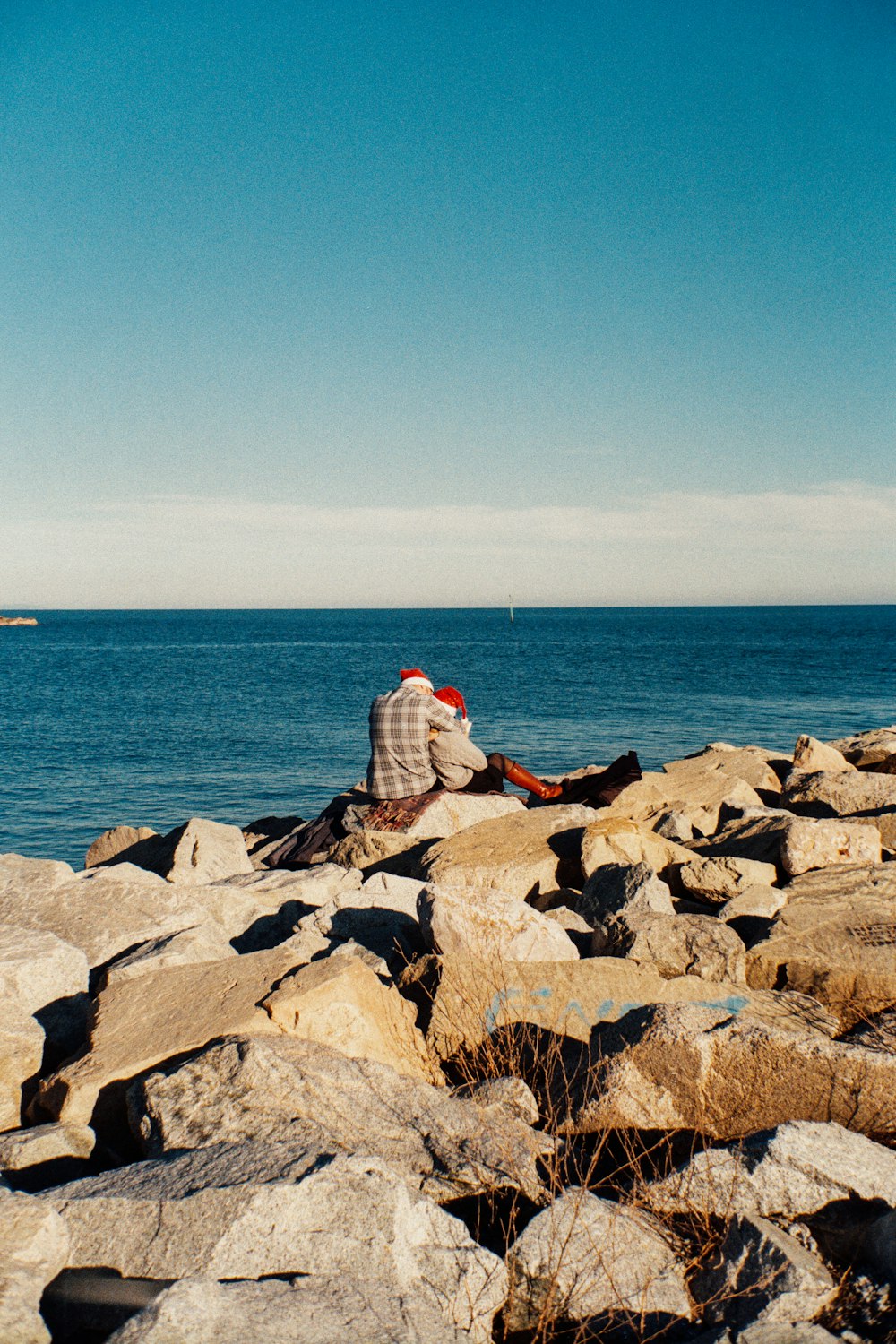 person in white and red shirt sitting on gray rock near body of water during daytime