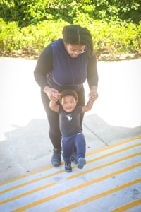 An adult is assisting a small child who is joyfully attempting to walk up a set of stairs. The adult, wearing dark clothing and glasses, holds the child's hands for support. The child is dressed in a dark-colored outfit and appears happy and confident while making steps. In the background, there is greenery with bright sunlight illuminating the scene.