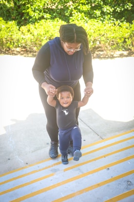 An adult is assisting a small child who is joyfully attempting to walk up a set of stairs. The adult, wearing dark clothing and glasses, holds the child's hands for support. The child is dressed in a dark-colored outfit and appears happy and confident while making steps. In the background, there is greenery with bright sunlight illuminating the scene.