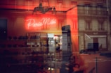 A warmly lit interior with a neon sign displaying the words 'Paris Miki'. Reflections of a city street and buildings appear on the glass, blending with the inside view where several pairs of sunglasses are displayed on shelves.