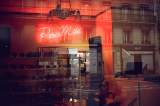 A warmly lit interior with a neon sign displaying the words 'Paris Miki'. Reflections of a city street and buildings appear on the glass, blending with the inside view where several pairs of sunglasses are displayed on shelves.