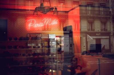 A warmly lit interior with a neon sign displaying the words 'Paris Miki'. Reflections of a city street and buildings appear on the glass, blending with the inside view where several pairs of sunglasses are displayed on shelves.