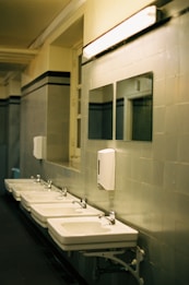 A row of white ceramic sinks with metal faucets is lined up against a tiled wall in a public restroom. Above the sinks are rectangular mirrors, and a fluorescent light fixture is mounted on the wall providing illumination. A soap dispenser is attached to the wall between the mirrors.