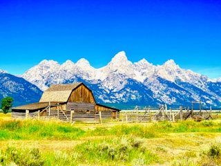 A rustic wooden barn stands in the foreground, surrounded by a wooden fence in a grassy field. Behind it, a range of snow-capped mountains stretches across the horizon under a clear blue sky.