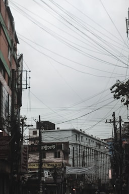 A city street with numerous overhead electrical wires crisscrossing against a cloudy sky. Buildings line both sides of the street, with signs in a foreign script visible on the facades. The scene conveys an urban environment with a slightly overcast atmosphere.