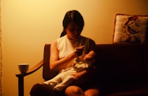 A woman is seated on a sofa, gently feeding a baby in her arms. The scene is softly lit, creating a warm, intimate atmosphere. A cup rests on the arm of the sofa, and a decorative pillow with bird designs is placed nearby.