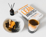 A book titled 'Lean Analytics' lies on a table next to a glass of coffee. There is a brown bottle labeled 'Rosemary' with diffuser sticks and a white bowl containing a dessert topped with caramel and accompanied by olives.