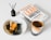A book titled 'Lean Analytics' lies on a table next to a glass of coffee. There is a brown bottle labeled 'Rosemary' with diffuser sticks and a white bowl containing a dessert topped with caramel and accompanied by olives.