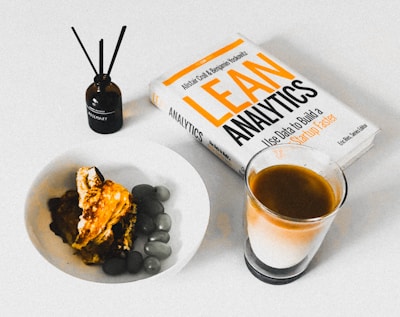 A book titled 'Lean Analytics' lies on a table next to a glass of coffee. There is a brown bottle labeled 'Rosemary' with diffuser sticks and a white bowl containing a dessert topped with caramel and accompanied by olives.