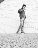 A man stands casually against a modern, geometric-patterned wall, holding sunglasses in one hand. He is dressed in a button-down shirt, jeans, and sneakers, with one hand in his pocket. The ground is textured, resembling stone or concrete.