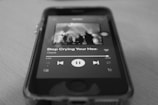 A close-up view of a smartphone displaying a music app interface with a song by Oasis titled 'Stop Crying Your Heart Out.' The screen shows playback controls and part of an album cover. The image is in black and white, focusing on the details of the screen and the phone's physical edges.