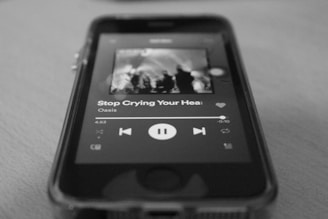 A close-up view of a smartphone displaying a music app interface with a song by Oasis titled 'Stop Crying Your Heart Out.' The screen shows playback controls and part of an album cover. The image is in black and white, focusing on the details of the screen and the phone's physical edges.
