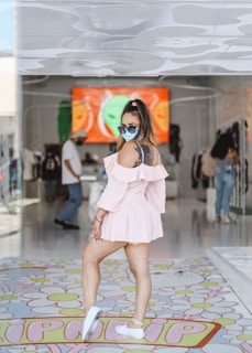 A person in a pink dress and blue sunglasses stands confidently in an indoor setting, which appears to be a modern store or boutique. The background includes a large, colorful digital screen, racks of clothing, and several other people. The floor has a colorful pattern with flowers and the word 'HIPPY' visible.