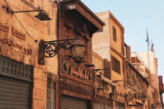 A street scene featuring an old building with Arabic script on the exterior walls. The architecture includes ornate wrought iron lamps and a wooden facade above a closed shop with a sign in Arabic. The walls are made of aged stone, contributing to a historic atmosphere.